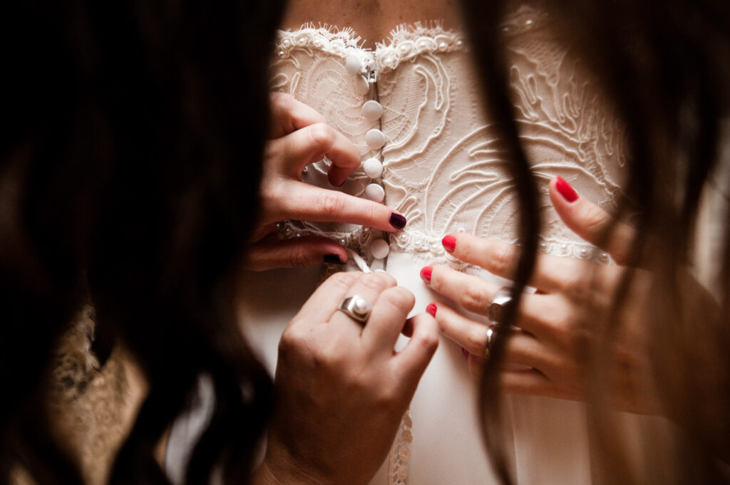 Dos mujeres, a los lados de la imagen, abrochan el vestido de novia de la que está en el centro.