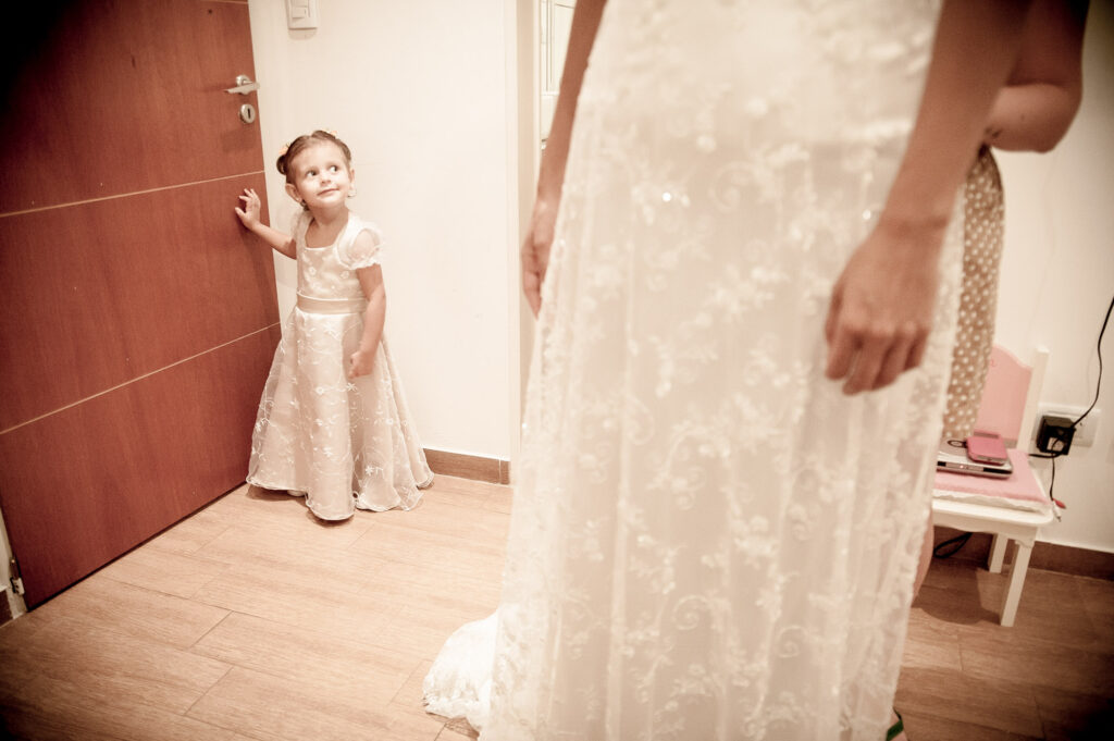 Una niña, en segundo plano en el margen superior izquierdo, ingresa a la habitación con un vestido blanco, donde su madre se está probando el vestido para su casamiento.
