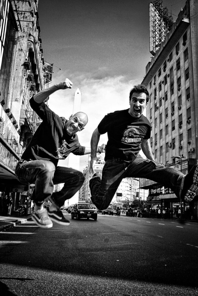 Dos actores de mayumana saltan en la Avenida Corrientes con el Obelisco de fondo.
